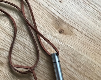 Op de afbeelding: Een zilveren metalen buisvormig hanger aan een bruin leren koord. De hanger is ongeveer 5 cm lang en heeft een gladde, gepolijste afwerking.