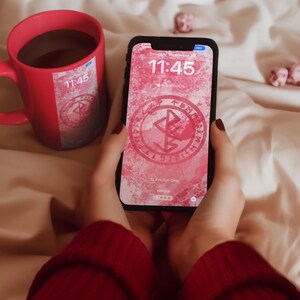 May include: A red mug with a black liquid inside and a phone with a pink and white background. The phone screen displays a clock with the time 11:45 and the date Saturday, September 30th. The phone also has a red symbol with a white outline on the screen.
