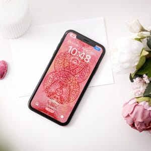 Peut inclure: Un smartphone avec un étui noir affiche un fond d'écran rose et rouge à motifs avec l'heure 10h48. Un bonbon rose en forme de cœur, une bougie blanche et des fleurs roses et blanches sont disposés sur une surface blanche.