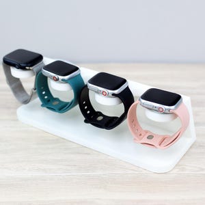 May include: A white stand with four slots for holding smartwatches. The stand has four smartwatches with different colored bands: black, teal, pink, and silver.