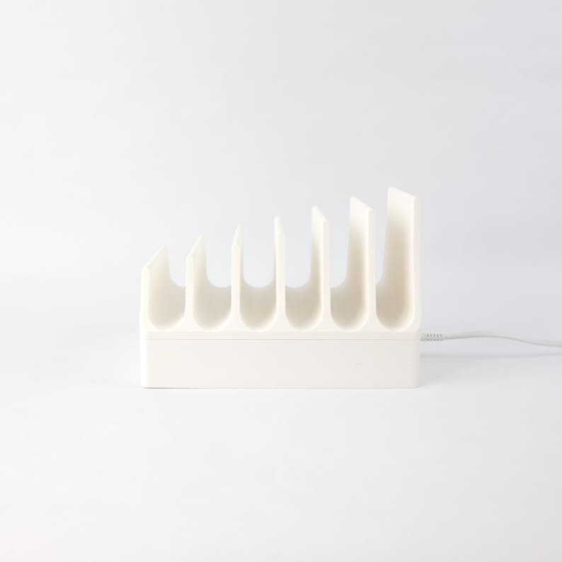 May include: White plastic charging station with six slots for charging devices. The station has a white cord attached to it.