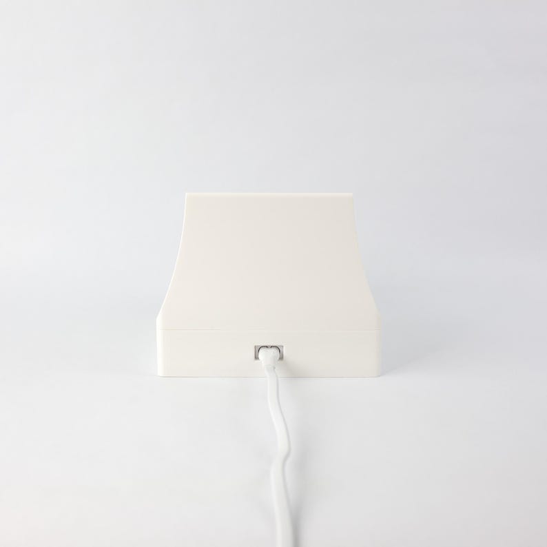 May include: A white, rectangular charging base with a white cord attached. The base has a slightly curved top and a flat bottom.
