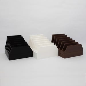 May include: Three different colored plastic storage organizers for video game cartridges. The organizers are black, white, and brown. Each organizer has six slots for cartridges.