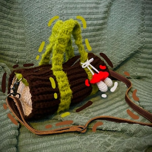 May include: A brown and green crocheted bag with a long brown leather strap. The bag is shaped like a log and has a mushroom design on the front.