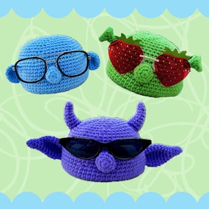 Puede incluir: Tres sombreros de ganchillo con diferentes rasgos faciales y gafas de sol. El primer sombrero es azul con gafas redondas, el segundo es verde con gafas de sol en forma de fresa y el tercero es morado con gafas de sol negras.