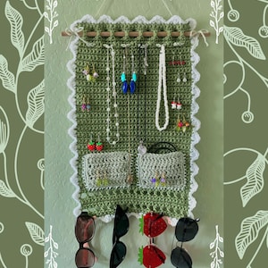 Puede incluir: Un organizador de pared de crochet verde con ribete blanco y bolsillos para guardar joyas y gafas de sol. El organizador tiene una varilla de madera en la parte superior para colgar collares y pendientes. Hay varios pares de gafas de sol colgando de la parte inferior del organizador.