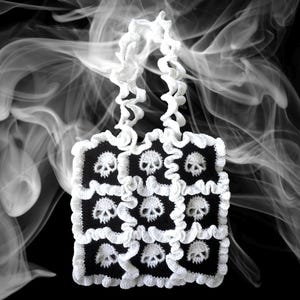 以下が含まれることがあります： 黒いドクロ柄の白いクロシェトートバッグ。バッグはフリルトリムとシングルストラップが付いています。