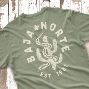 May include: Sage green t-shirt with a circular graphic that reads "BAJA NORTE EST. 1931." The graphic features a stylized cactus and snake design in a cream color. The shirt is made of a soft material.