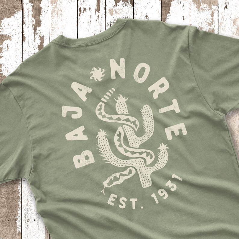 May include: Sage green t-shirt with a circular graphic that reads "BAJA NORTE EST. 1931." The graphic features a stylized cactus and snake design in a cream color. The shirt is made of a soft material.