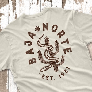 May include: Cream-colored t-shirt with a brown graphic design on the back. The design features a cactus, a snake, and the words "BAJA NORTE EST. 1931". The design has a distressed, vintage look.