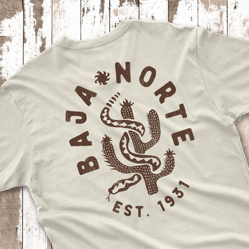 May include: Cream-colored t-shirt with a brown graphic design on the back. The design features a cactus, a snake, and the words "BAJA NORTE EST. 1931". The design has a distressed, vintage look.