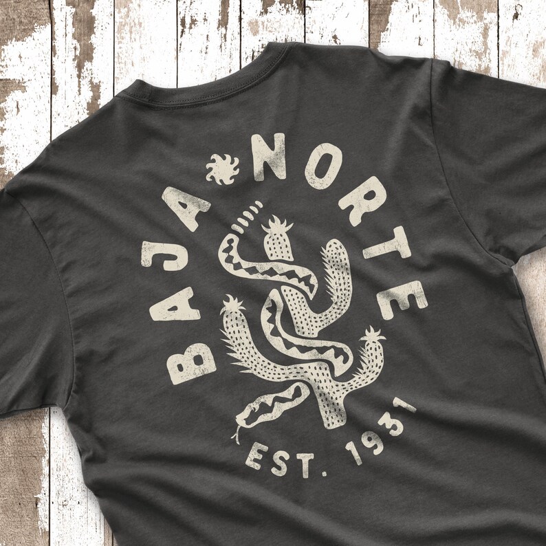 May include: Dark gray t-shirt with a distressed white graphic. The graphic features a stylized cactus intertwined with a snake, surrounded by the words "BAJA NORTE EST. 1931". The design has a vintage, worn look.