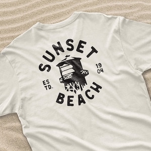 May include: Cream-colored t-shirt featuring a black graphic design on the back. The design includes the words "SUNSET BEACH" in a circular arrangement around a stylized lifeguard tower. The text "ES TD. 19 04" is also included.