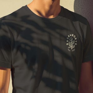 May include: Dark gray t-shirt with a circular graphic featuring a stylized cactus and the words "BAJA NORTE" and "EST. 1931". The design is in a lighter shade, creating a subtle contrast against the shirt's color.