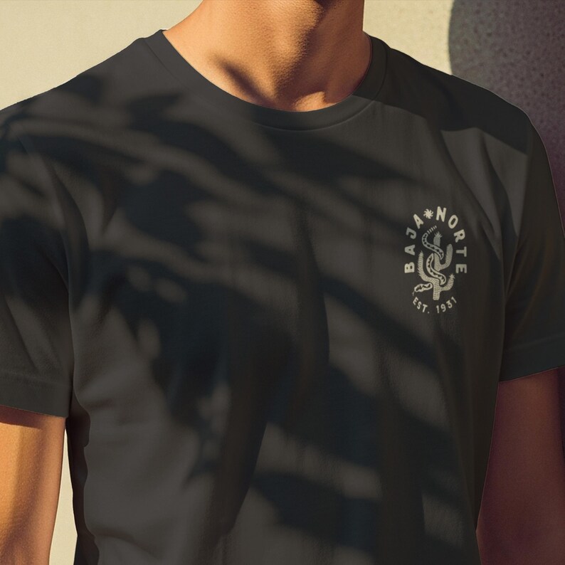 May include: Dark gray t-shirt with a circular graphic featuring a stylized cactus and the words "BAJA NORTE" and "EST. 1931". The design is in a lighter shade, creating a subtle contrast against the shirt's color.