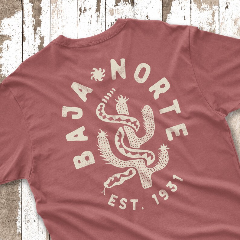 May include: A dusty rose t-shirt with a cream-colored graphic on the back. The graphic features a cactus and snake design with the words "BAJA NORTE EST. 1931" in a circular arrangement. The shirt is made of soft material.