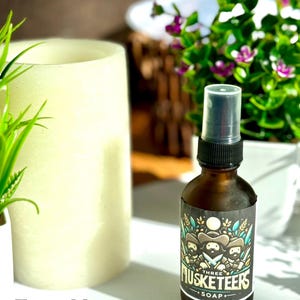 May include: A brown glass bottle with a black spray top and a label that says "Three Musketeers Soap" with a cartoon image of three musketeers. The bottle is sitting on a white surface.