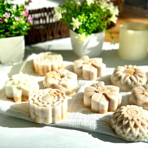 May include: A collection of eight handmade soap bars in various floral designs. The soaps are white with a light brown swirl and are arranged on a white towel.