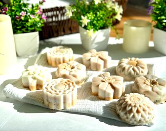 May include: A collection of eight handmade soap bars in various floral designs. The soaps are white with a light brown swirl and are arranged on a white towel.