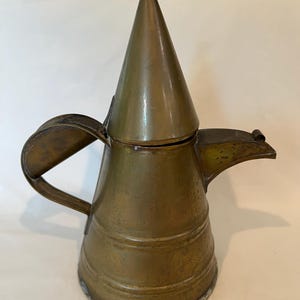 May include: An antique brass-coloured metal pitcher with a conical lid and a small handle. The pitcher has a spout and a larger handle on the side. The metal has a tarnished appearance, suggesting age. The pitcher is likely for decorative use.