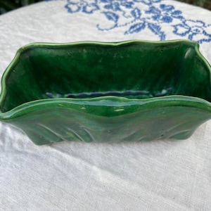 May include: A rectangular, green ceramic planter with a wavy, leaf-like design. The planter has a glossy finish and sits on a white surface with a blue floral pattern.