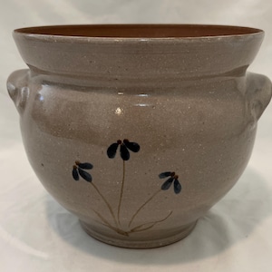 May include: A handcrafted, speckled stoneware planter with a wide opening and two side handles. The exterior is a muted gray-brown, adorned with hand-painted blue flower accents. The interior rim is a darker brown.