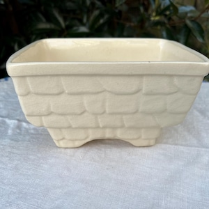 Könnte beinhalten: Ein weißer Keramiktopf mit einem strukturierten Design, das an eine Backsteinmauer erinnert. Der Topf hat vier kleine Füße und ist ideal für kleine Pflanzen.