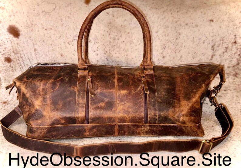 May include: A brown leather duffel bag with two top handles and a detachable shoulder strap. The bag has two zipped pockets on the front and a distressed finish. The text "HydeObsession.Square.Site" is at the bottom.