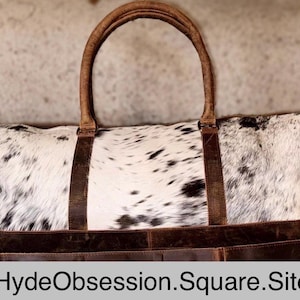 May include: A large, rectangular duffel bag with a cowhide pattern in black and white. The bag features brown leather handles, straps, and trim. The bag has a shoulder strap and the text "HydeObsession.Square.Site" is visible.
