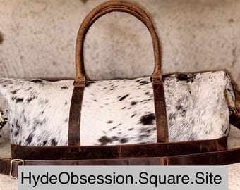May include: A large, rectangular duffel bag with a cowhide pattern in black and white. The bag features brown leather handles, straps, and trim. The bag has a shoulder strap and the text "HydeObsession.Square.Site" is visible.