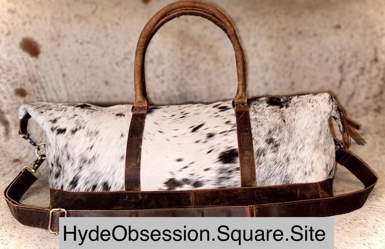May include: A large, rectangular duffel bag with a cowhide pattern in black and white. The bag features brown leather handles, straps, and trim. The bag has a shoulder strap and the text "HydeObsession.Square.Site" is visible.