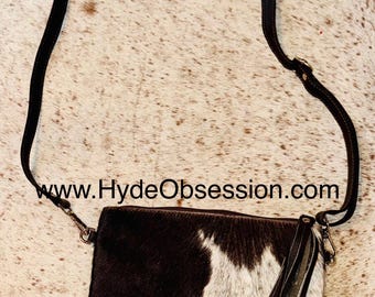 May include: A black and white cowhide crossbody bag with a brown leather strap and a long tassel. The bag has a zipper closure and a small metal buckle on the strap. The website address "www.HydeObsession.com" is printed on the bag.