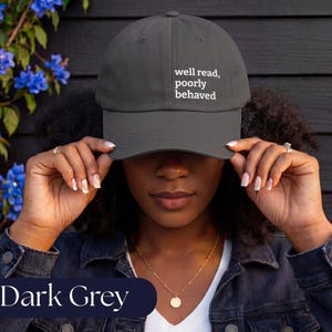 May include: A dark grey baseball cap featuring the white text "well read, poorly behaved" embroidered on the front panel. The cap has a structured crown, a curved brim, and a classic six-panel design with a button on top and stitched eyelets.