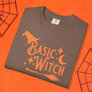 May include: A brown t-shirt with the words "BASIC WITCH" in orange. The design includes a witch hat, stars, a crescent moon, and a broom. The shirt is displayed on an orange background with spiderweb decorations.
