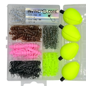 May include: A clear plastic box containing a variety of fishing lures. The box has six compartments, each filled with a different type of lure. The lures are in various colors, including brown, pink, black, and green. The box also includes four yellow foam bobbers. The box has the text "INVINCI CORE" printed on the top.