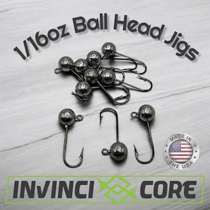 May include: A collection of ball head jigs with curved hooks. The image displays several jigs with a 1/16oz weight, arranged on a light wood-grain surface. The text "1/16oz Ball Head Jigs" is arched above the jigs. The image also includes a "Made in the USA" sticker and the "INVINCI CORE" logo.