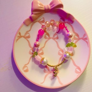 May include: A beaded bracelet with pink, clear, and gold beads, including a large pearl and a gold charm, rests on a pink and white decorative plate with a bow design. The plate has a bow-shaped handle.