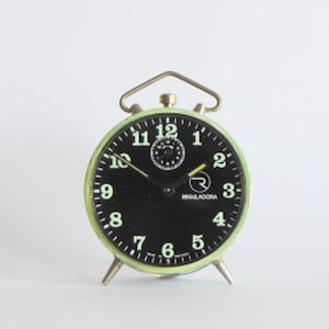 Puede incluir: Un reloj despertador vintage verde con una esfera negra y números blancos. El reloj tiene un marco de metal y una campana en la parte superior. La esfera del reloj tiene un pequeño dial de segundero y la palabra "REGALADORA" impresa en él.