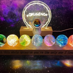 Könnte beinhalten: Sieben schimmernde Glaskugeln mit galaktischen Mustern sind auf einer Holzbasis mit dem Text "In diesem ganzen Universum bist du der Einzige, den ich liebe." angeordnet. Eine leuchtende Astronautenfigur steht vor einem runden, leuchtenden Schild mit dem Text "OOMUGENOO."
