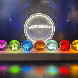 Op de afbeelding: Een display van zeven verlichte glazen bollen in verschillende kleuren, elk met een uniek geëtst ontwerp. De bollen zijn gerangschikt op een houten basis, met een gloeiend cirkelvormig bord met de tekst "OMUGENDO" op de achtergrond.
