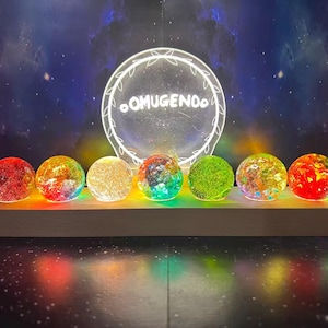 May include: A decorative light display with seven colourful, glowing orbs on a wooden base. A circular sign with the text "OMUGENDO" is illuminated above the orbs. The background is a starry night sky.