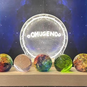 Può includere: Esposizione di sette globi sferici colorati su una base di legno. I globi variano di colore, tra cui rosso, giallo, verde e trasparente, con motivi interni. Un cartello circolare luminoso con la scritta "OMUGENDO" è sullo sfondo.