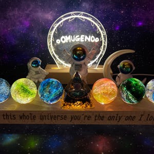 May include: A set of seven glass orbs with galaxy-like patterns, arranged on a wooden base with the text "In this whole universe you're the only one I love."  Two astronaut figurines and a light-up sign with the text "OOMUGENOO" are in the background.