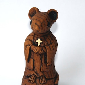 Pode incluir: Uma figurinha de cerâmica marrom de um rato vestido de monge, segurando uma cruz dourada em suas mãos.