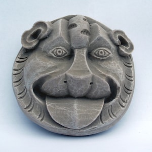 Pode incluir: Uma escultura de pedra cinzenta de uma gárgola com a boca aberta e a língua de fora. A gárgola tem um rosto detalhado com olhos, nariz e barba.