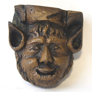 May include: A carved wooden face with large ears, a wide smile, and a textured surface. The face is brown and has a whimsical expression.