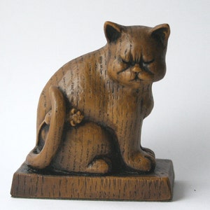 May include: A wooden figurine of a cat sitting with its tail curled around its body. The cat has a grumpy expression and is painted in a brown wood tone.