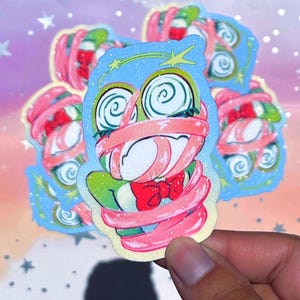 May include: A glittery die-cut sticker featuring a green frog character with swirling eyes, wrapped in pink bandages, and wearing a red bow tie. The character is set against a light blue background with yellow star accents and a sparkling finish.