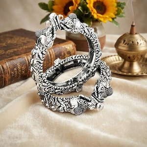 Puede incluir: Dos brazaletes de metal plateado con intrincados patrones florales en relieve y motivos prominentes de cabezas de elefante en el área del cierre. Las pulseras tienen un acabado oscuro y oxidado en las hendiduras para resaltar el metal.