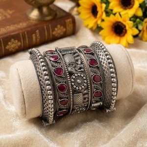 Op de afbeelding: Een brede zilverkleurige armband met ingewikkeld metaalwerk, inclusief reliëfmotieven van olifanten en cirkelvormige patronen. Het ontwerp is geaccentueerd met meerdere druppelvormige dieprode stenen in het gestructureerde metalen frame.